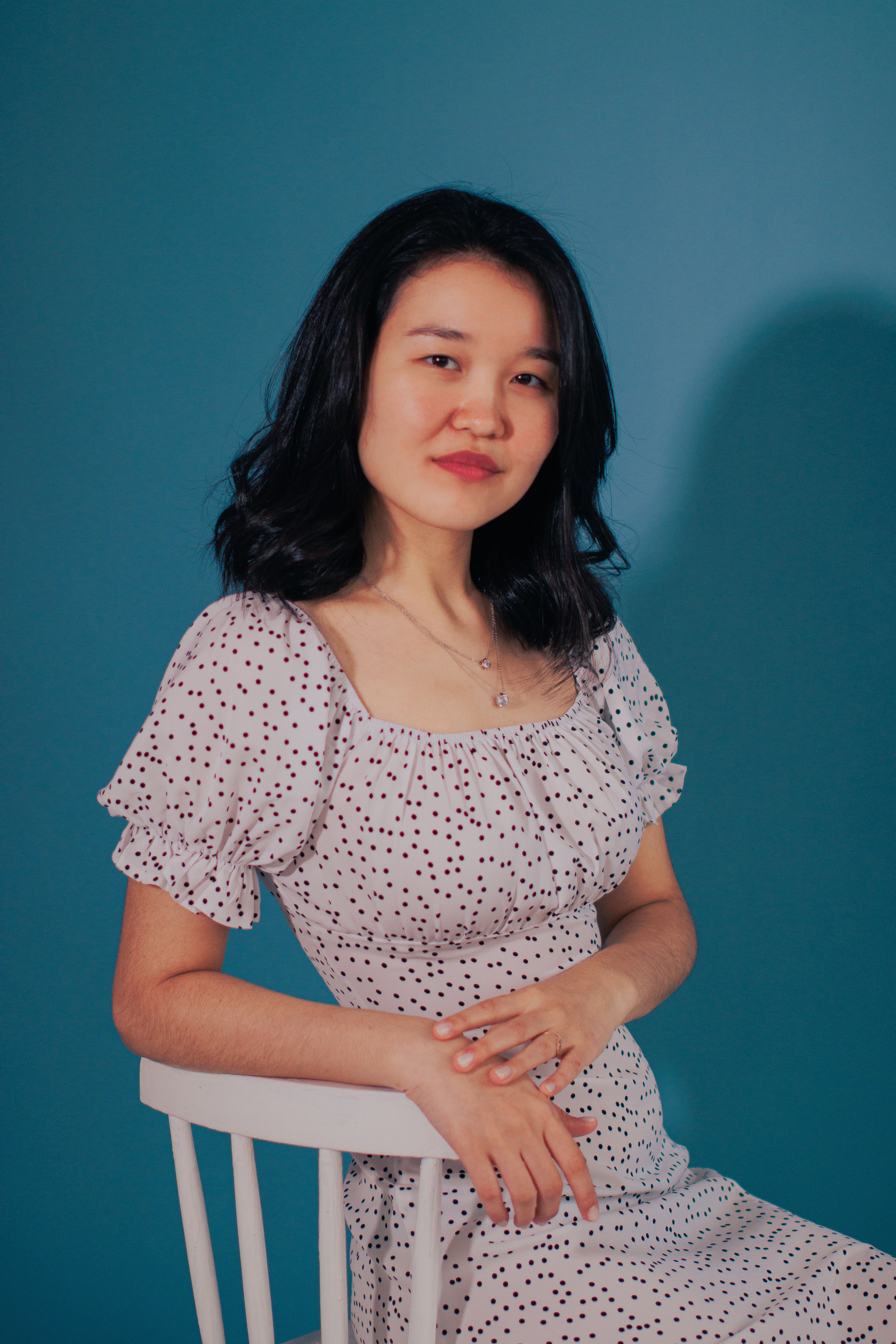 Elegant portrait of a woman sitting on a chair in a white spotted dress against a blue background.