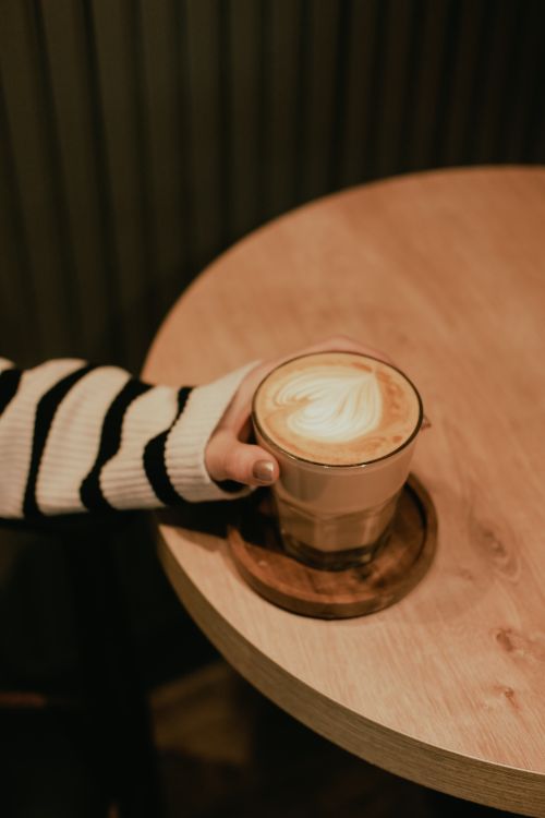 Free A person holding a cappuccino with latte art on a wooden table in a cozy cafe setting. Stock Photo