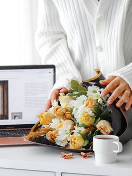 A woman in a white sweater arranges a bouquet on a desk with a laptop and coffee.