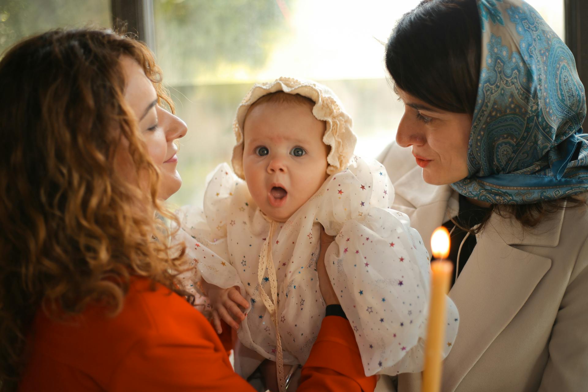 A joyful baby held by two women during a christening ceremony with candlelight.