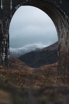 Scenic view through Glenfinnan Viaduct arch with snow-capped mountains and cloudy sky.