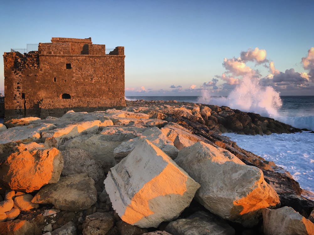 Ancient stone fortress on rocky coastline with waves crashing against shore at sunset.