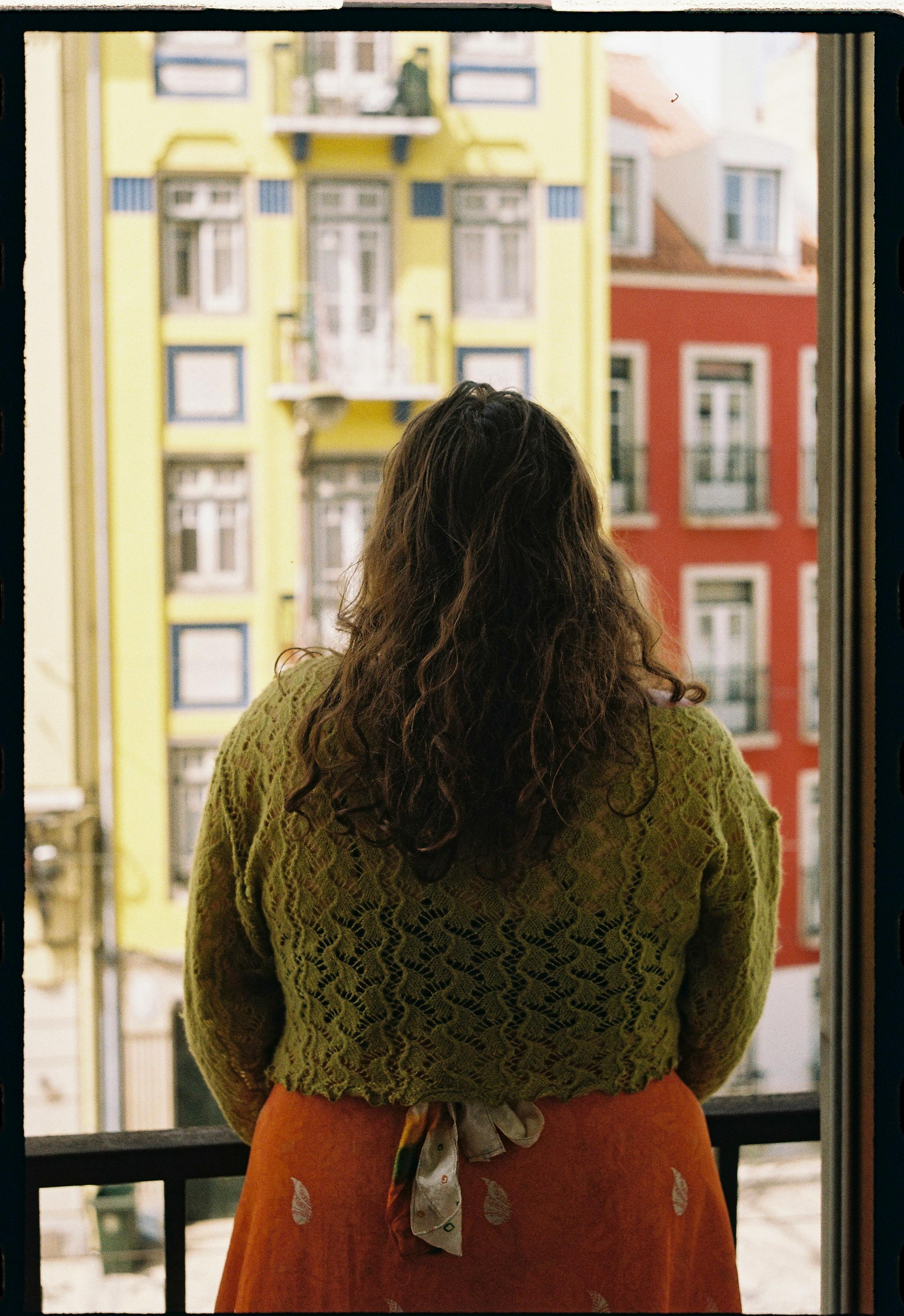 Woman with long brown hair in Lisbon gazing at colorful buildings from a balcony.