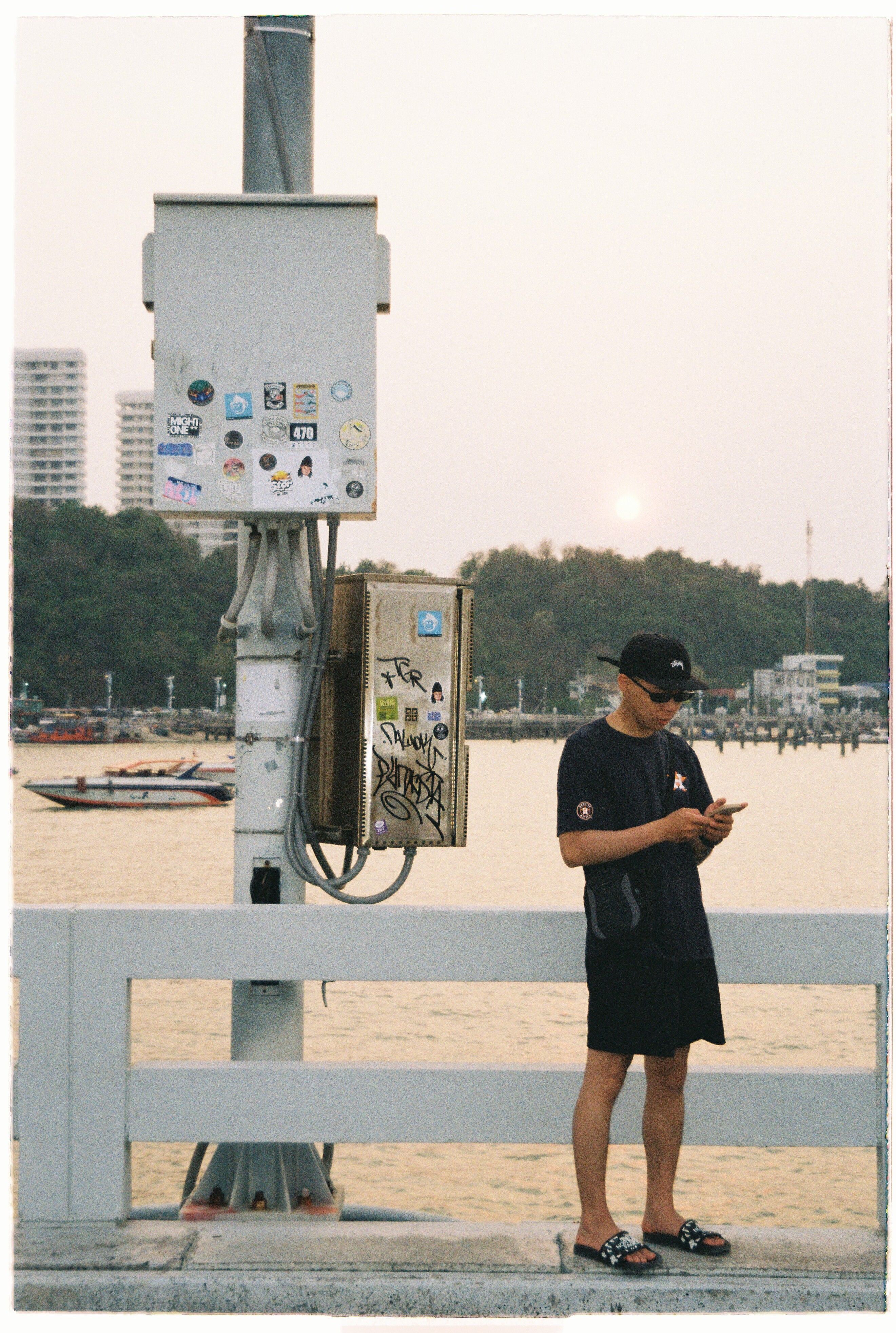 A man stands on a pier by the harbor using a phone at sunset, with a scenic cityscape backdrop.