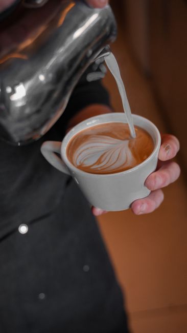 A stainless steel pitcher pouring steamed milk into a glass of espresso, creating a thin white ring of foam on top.