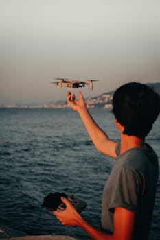 A young man controls a drone at sunset near the ocean, capturing a scenic view.