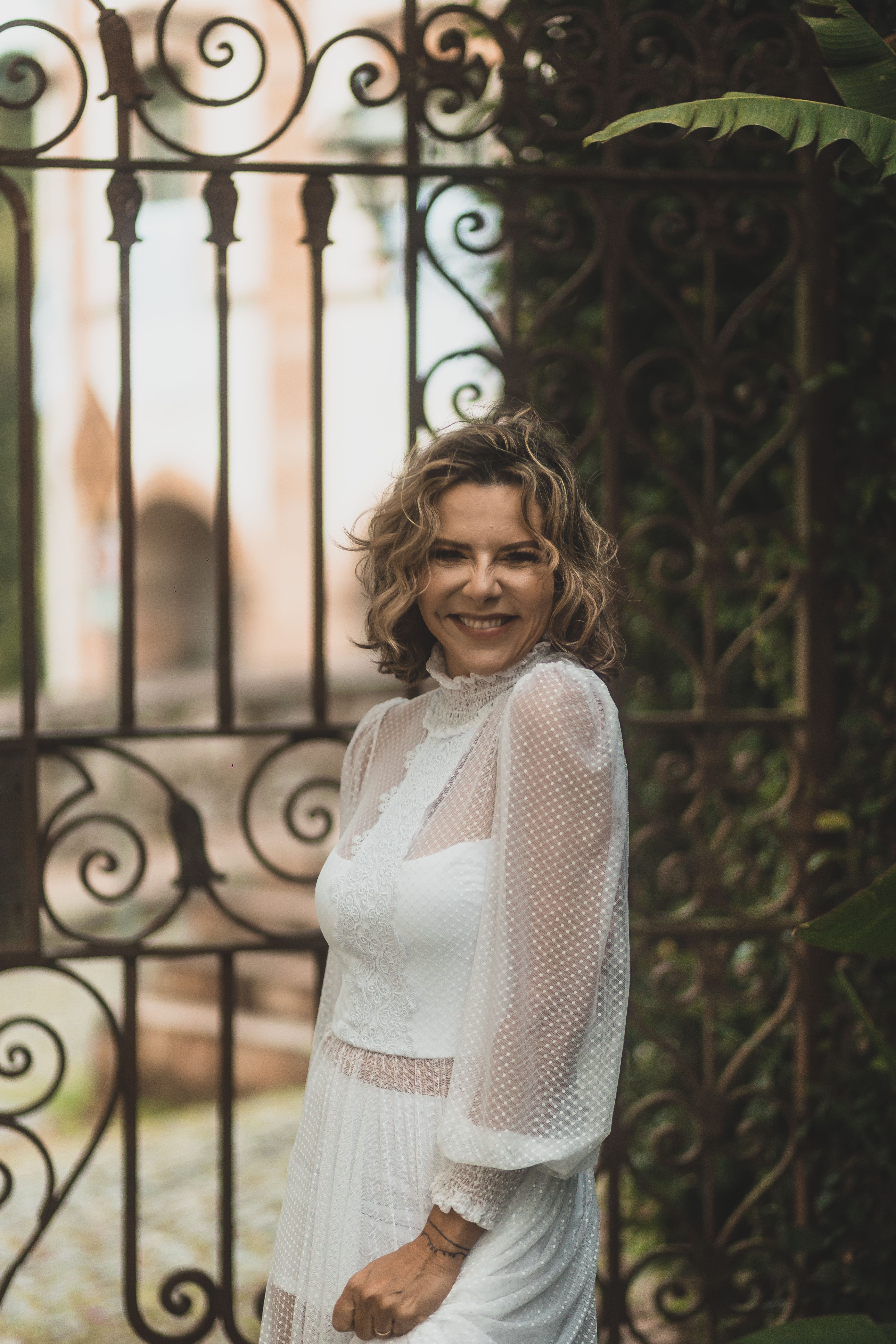A woman in a beautiful lace wedding dress smiling and posing outside near an ornate iron gate.