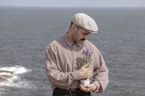 Free Man in vintage attire holding flowers by the seaside, exuding a nostalgic charm. Stock Photo