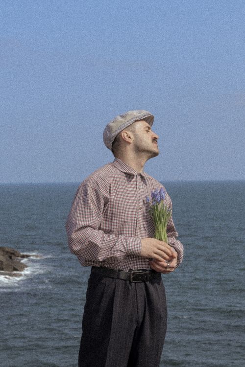 Free Man in classic attire holding flowers by the seashore, evoking a retro feel. Stock Photo