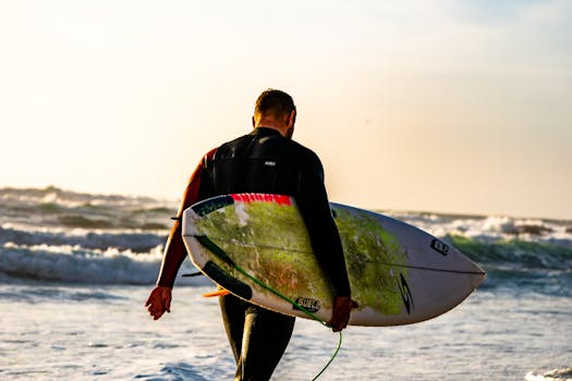 A surfer in wetsuit carrying a surfboard on a sunlit beach at sunset.
