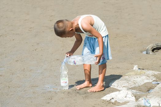 Young boy pours water from a plastic bottle on sandy beach during sunny day, symbolizing sustainability.