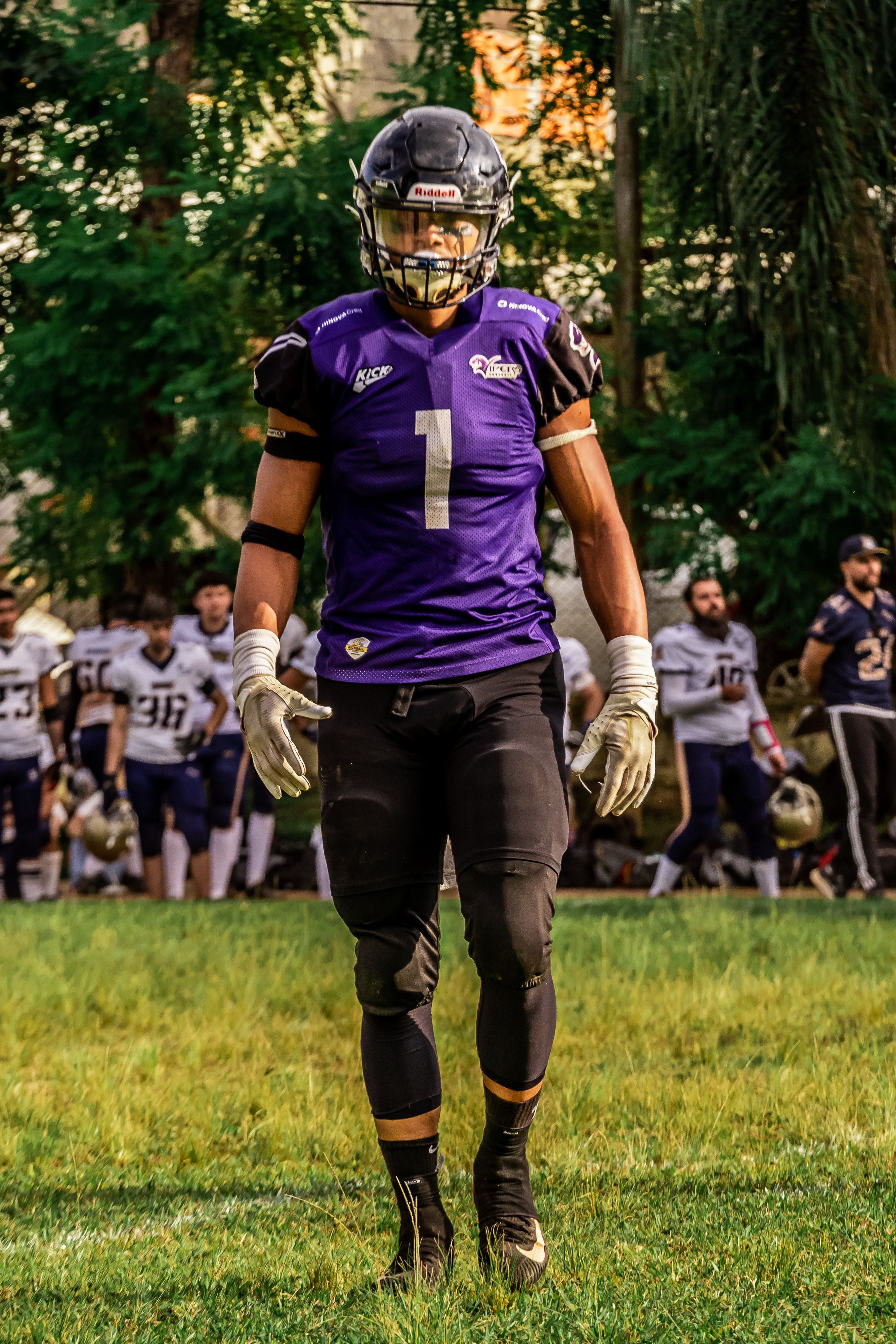 American football player in gear standing on field during a daytime match with team in the background.
