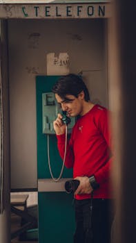 Man in a red shirt speaks on a vintage telephone in a booth, captured in a nostalgic setting.
