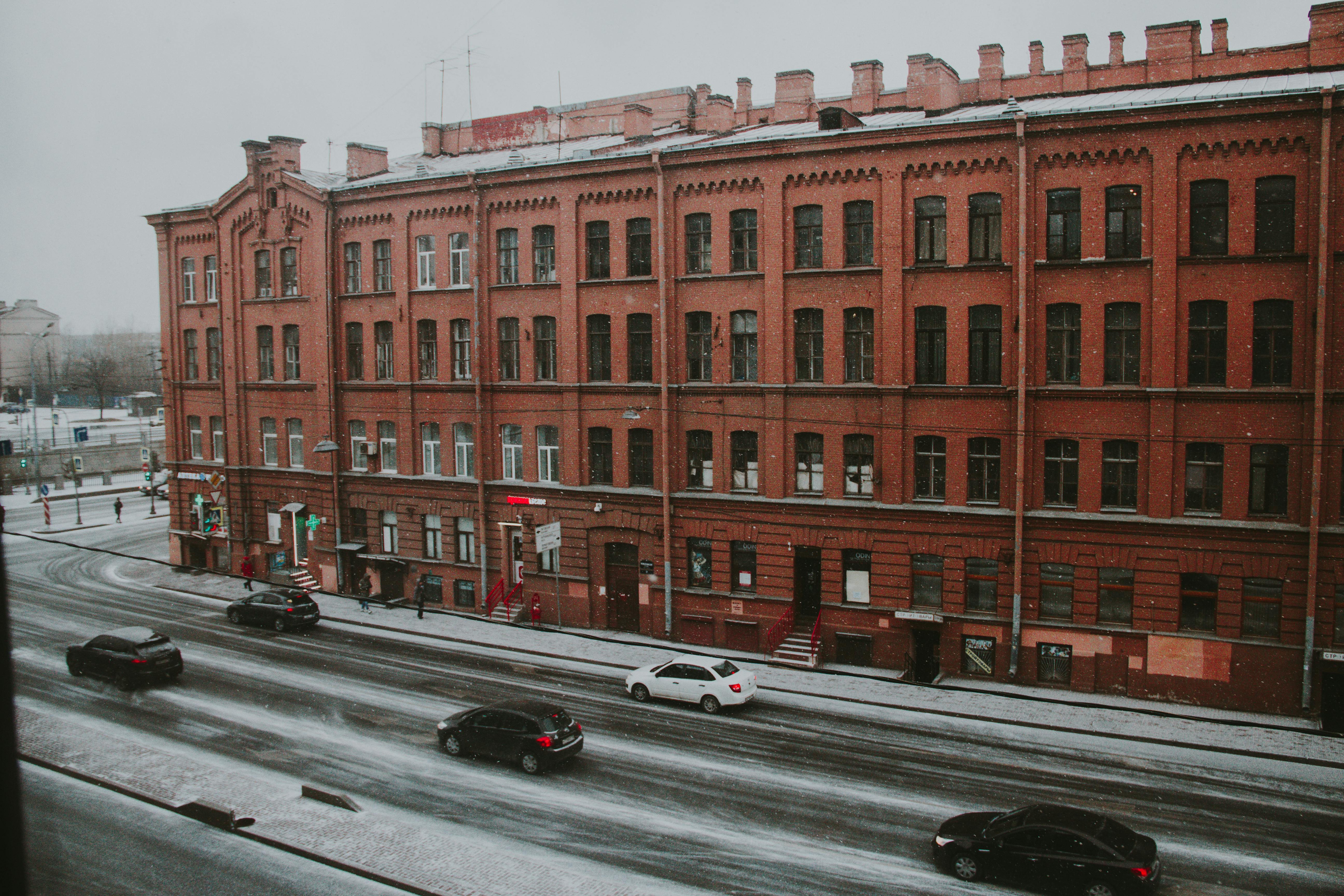 Free A snow-covered street with a historic red brick building and cars passing by, creating a winter urban scene. Stock Photo