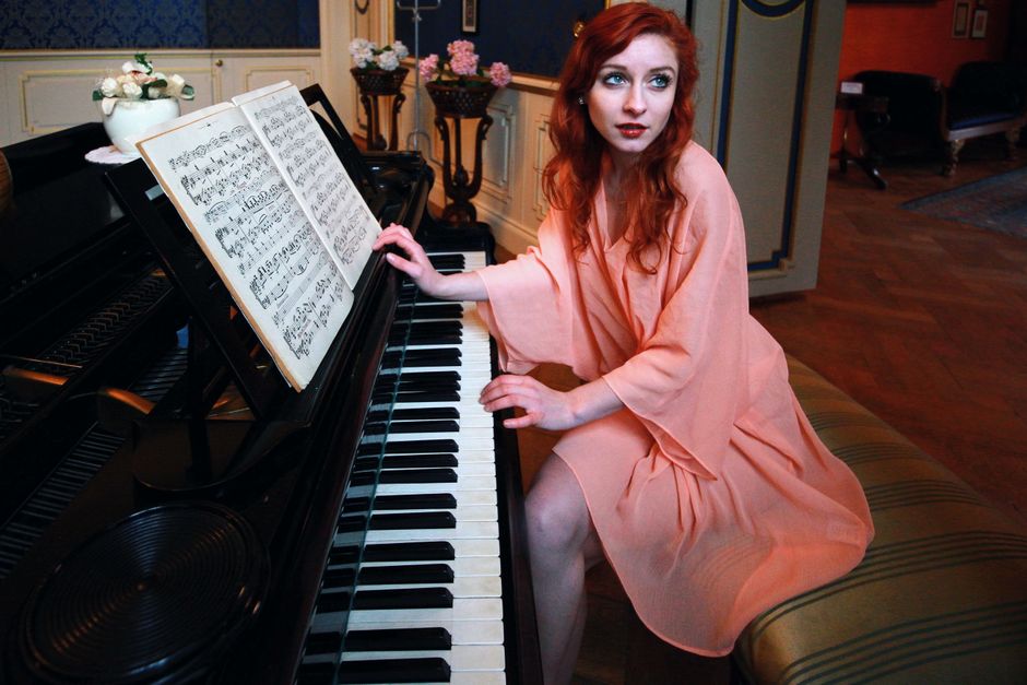 Red-haired woman in elegant attire plays piano in a luxurious room with flower decor and music sheets.