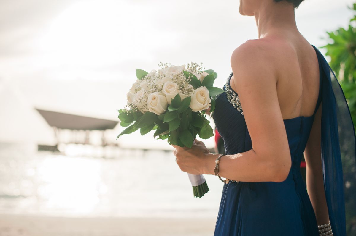 A woman in an elegant dress holds a bridal bouquet near the ocean at sunset.