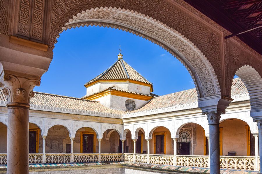 A Moorish arch framing a tiled pavilion roof and white arcades under a clear sky in Seville