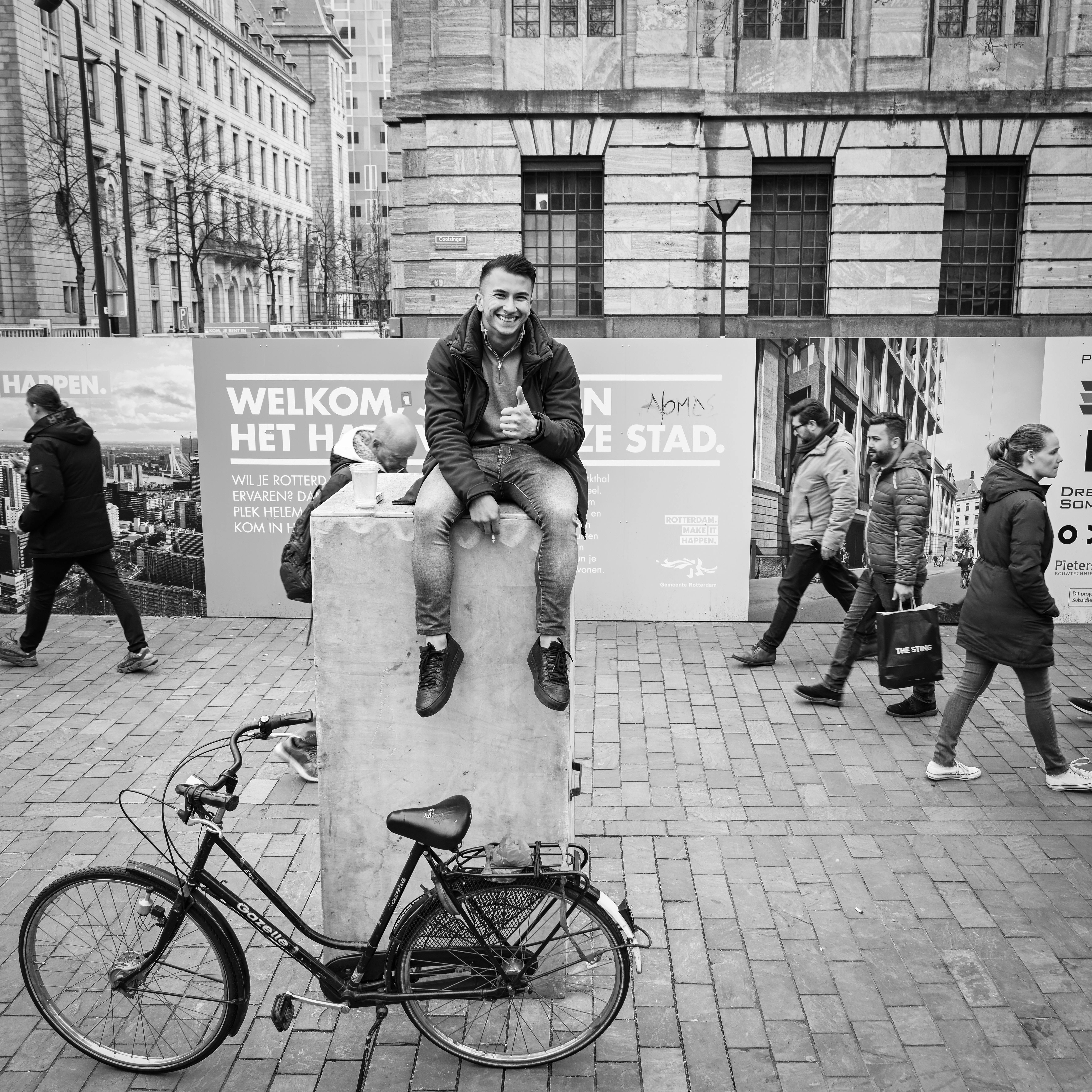 A man sitting on a stone, giving a thumbs up, in a bustling city street.