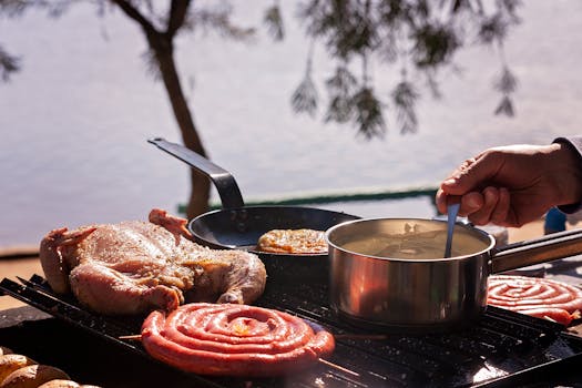 A scenic outdoor barbecue with chicken and sausage cooking over a grill, lake in the background.