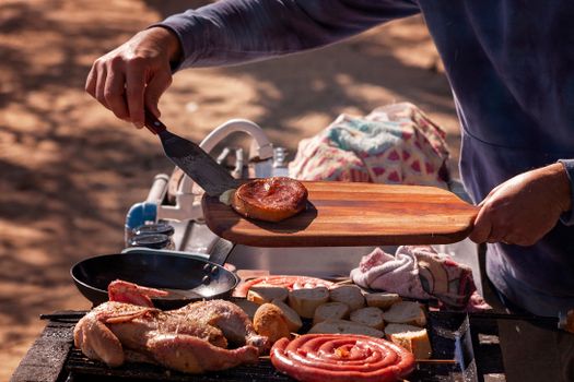 Close-up of delicious grilled chicken and sausages at an outdoor barbecue using a cutting board and spatula.