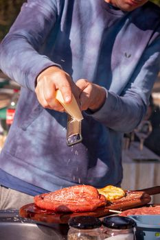 Man in blue sweatshirt grating cheese on sausages at a barbecue under sunlight, showcasing outdoor cooking.