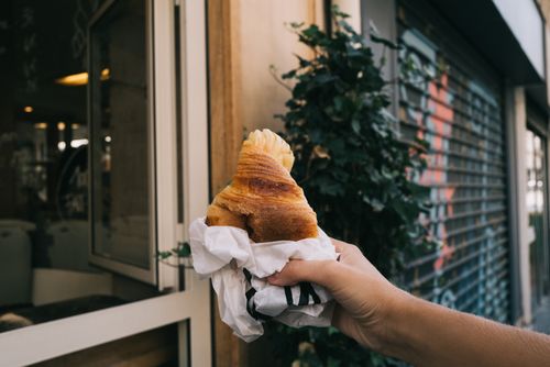 Free A person holding a croissant wrapped in paper in front of a café window. Urban vibe. Stock Photo
