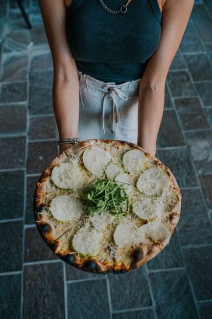 A woman holds a freshly baked pizza topped with cheese outdoors on a pavement.