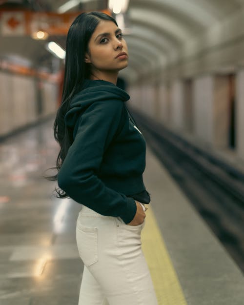 A woman in white jeans and a black hoodie standing on a subway platform
