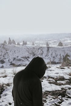 A person in a hoodie stands in a snowy winter landscape with unique rock formations.