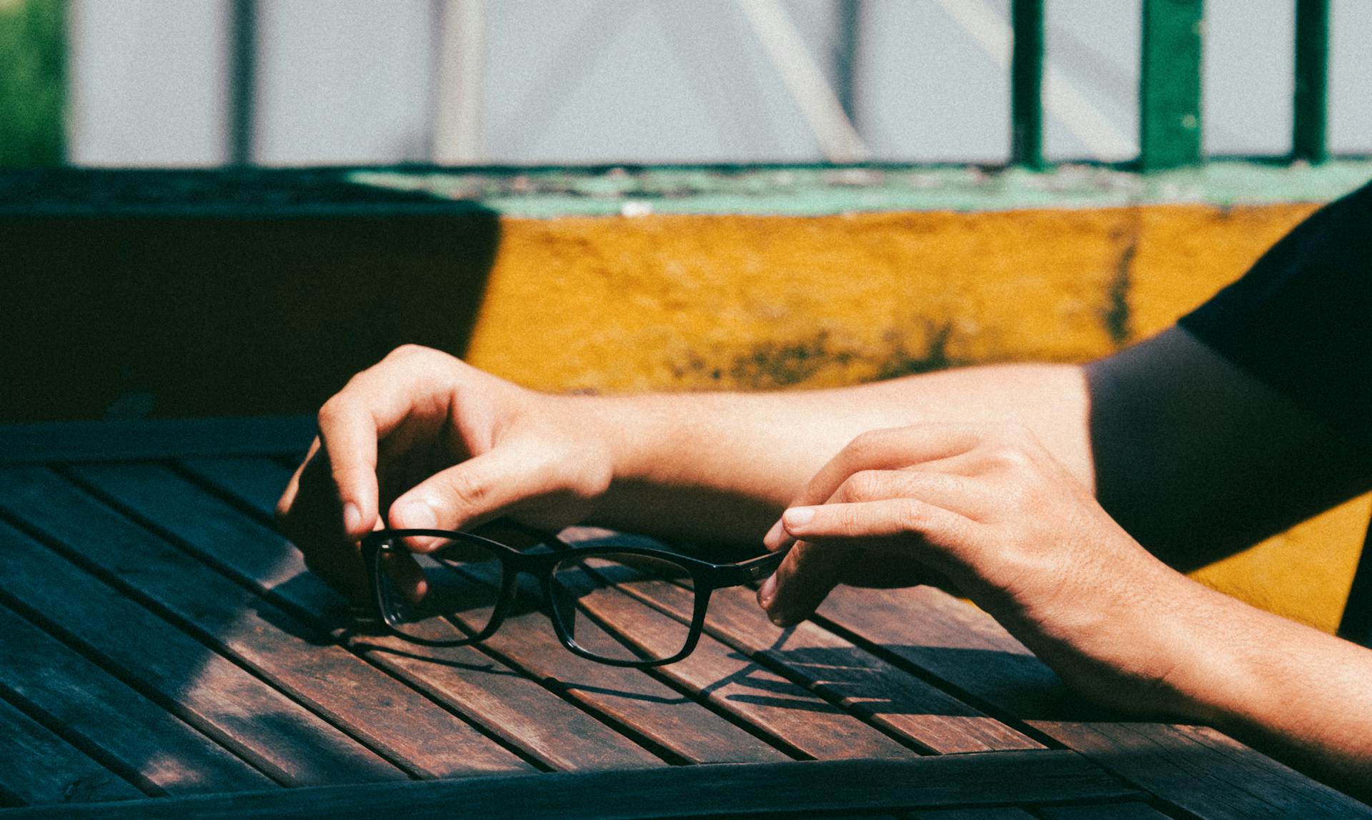 Close-up of hands holding eyeglasses on a wooden table outdoors in daylight.