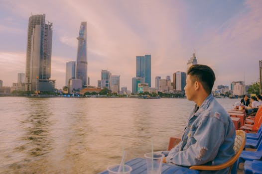 一名穿着牛仔夹克的男子坐在河边，欣赏日落时分的现代城市天际线