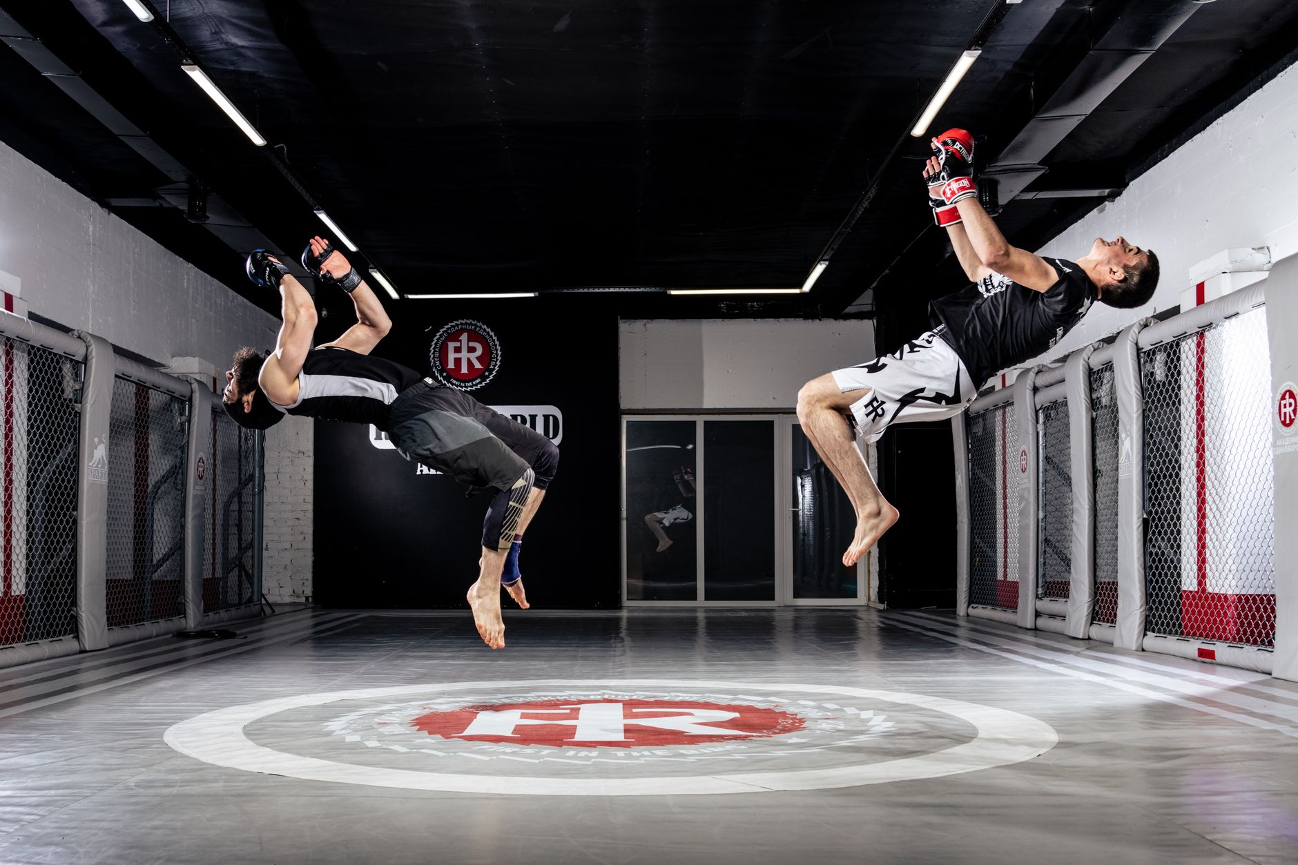 Two athletes perform dynamic flips in a martial arts training gym.