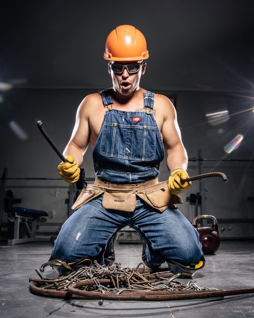 Free Muscular worker in hard hat and denim overalls poses with rebar and chains indoors. Stock Photo