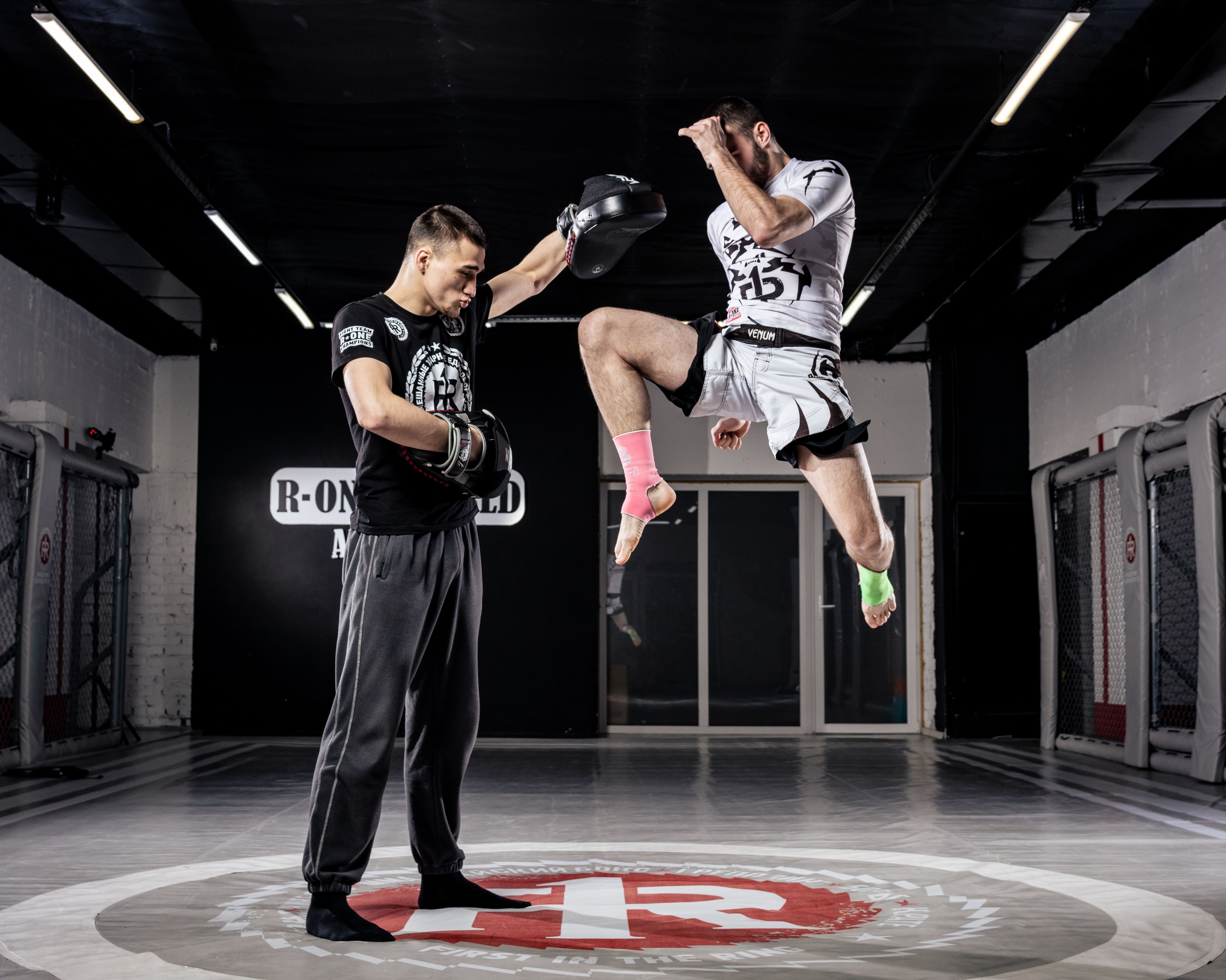 Two men practicing martial arts in a gym, showcasing a high kick and focus on training.