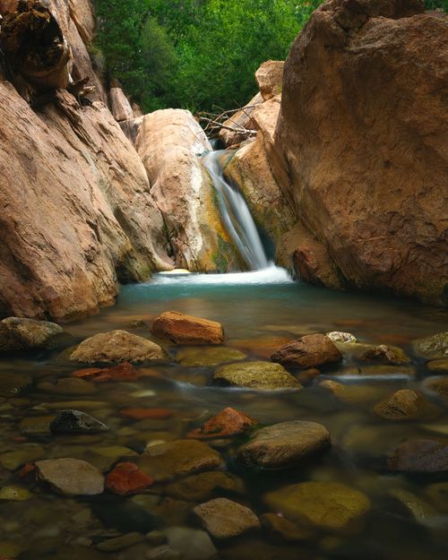 Free Tranquil waterfall cascading through a rocky canyon surrounded by lush greenery and smooth pebbles. Stock Photo