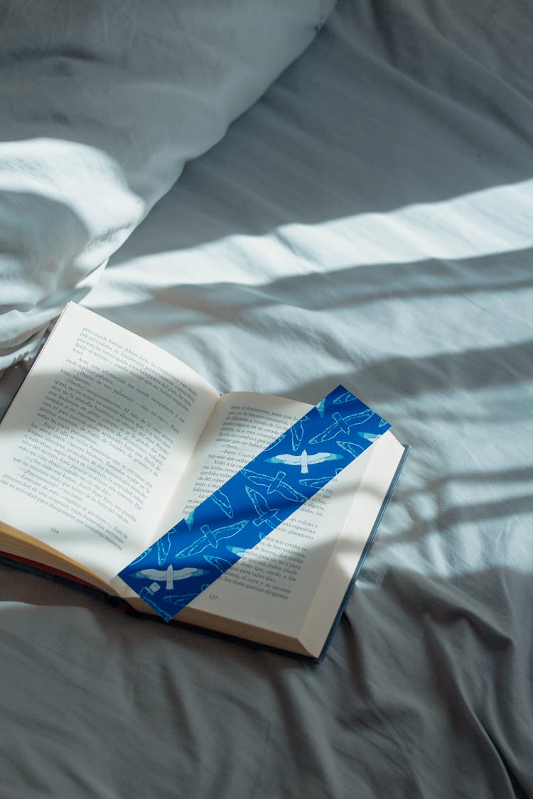 Photo Of An Open Book With A Blue Bookmark Lying On The Bed