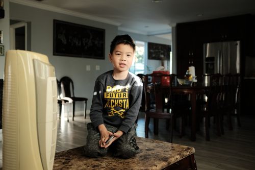 Free A young boy kneeling on a table in a sunlit living room, wearing a shirt with printed text. Stock Photo