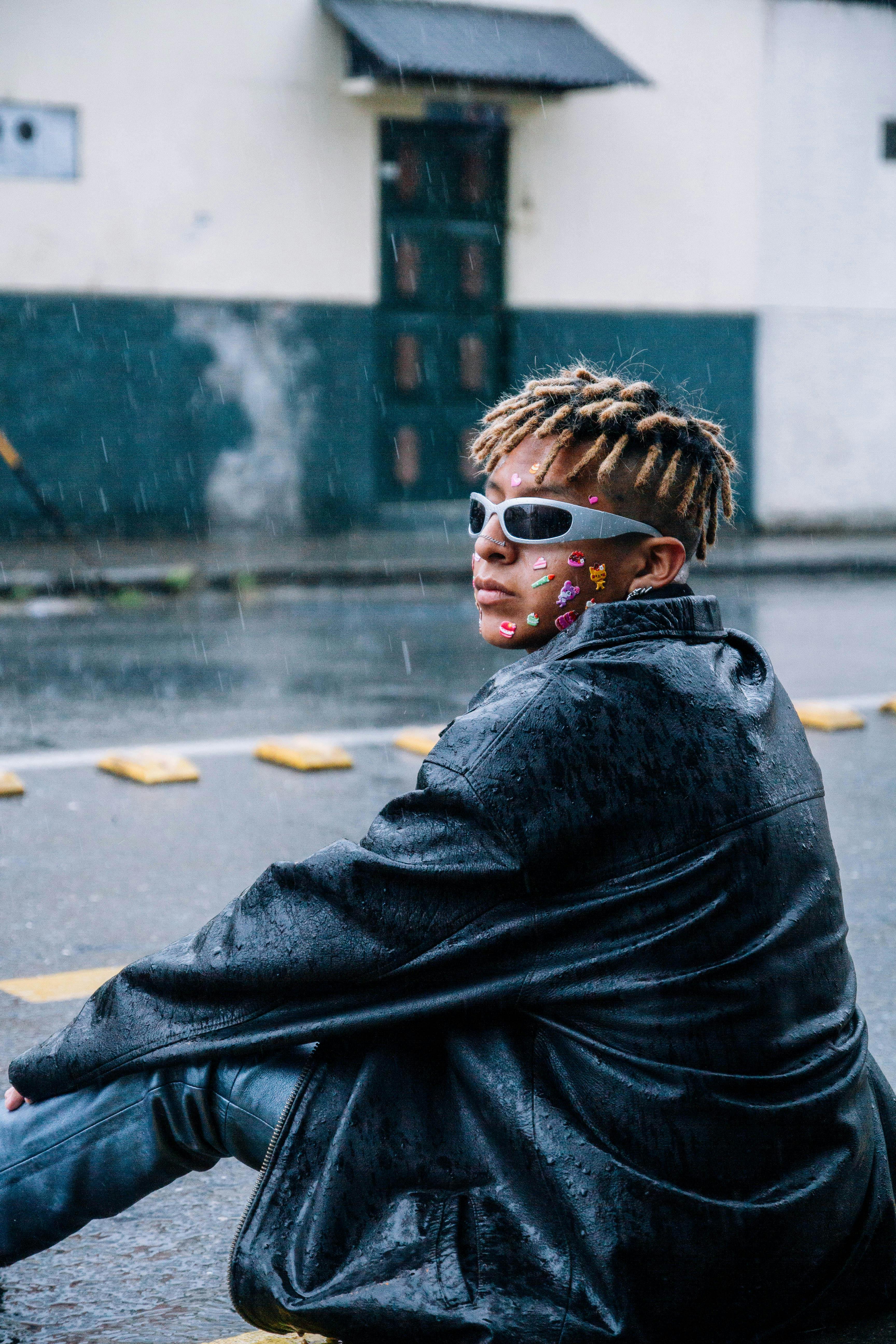 Fashionable teenager in sunglasses and leather jacket on a rainy street, featuring facial stickers.