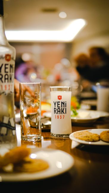 A glass of clear raki turning cloudy white as water is poured in, served alongside small meze plates of white cheese and melon.