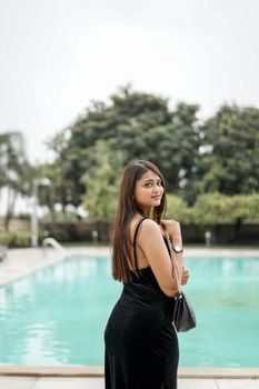 A stylish woman poses in a black dress by a poolside, exuding summer elegance.