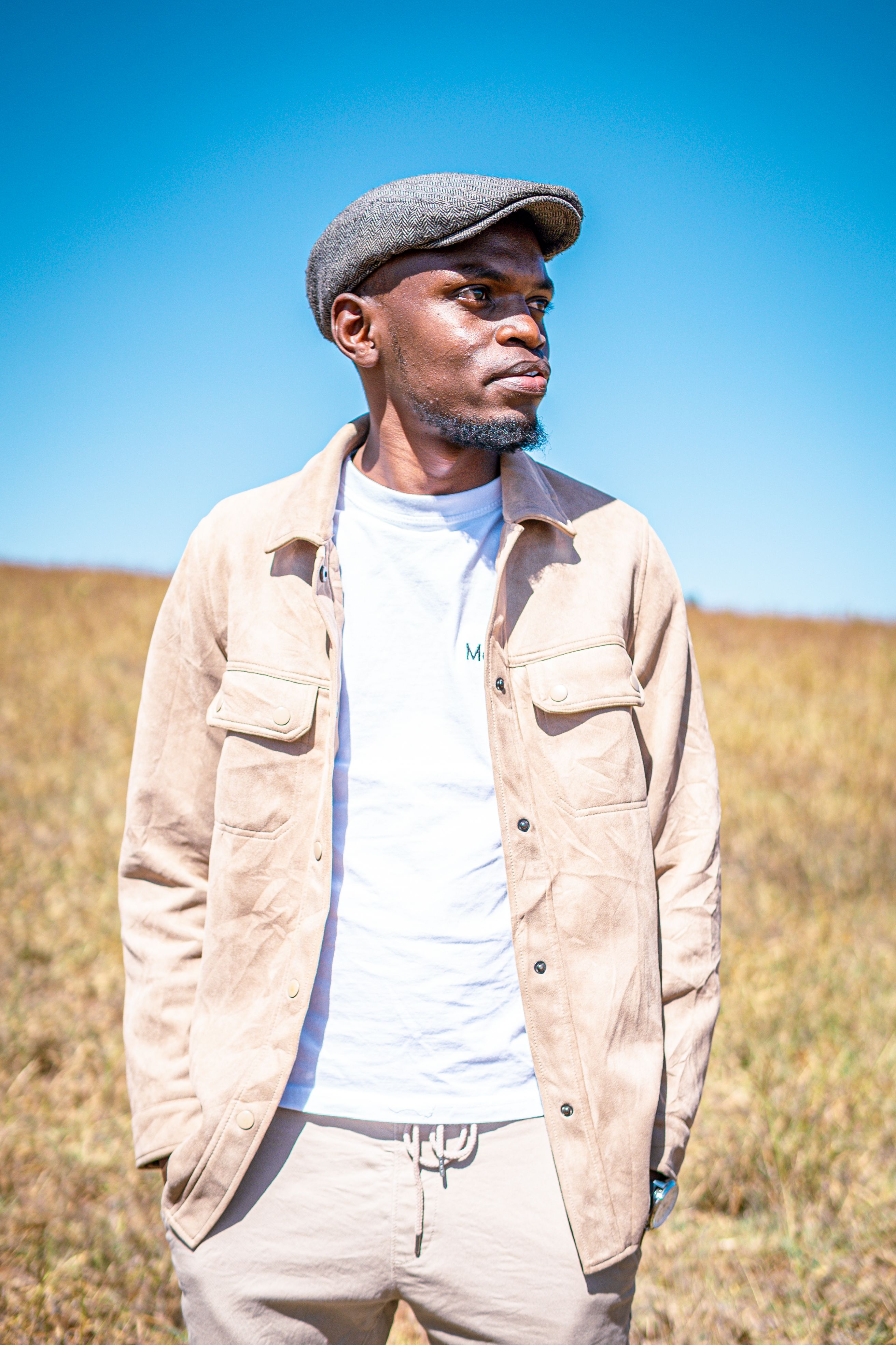 Portrait of a stylish man wearing a hat and jacket, posing outdoors in a sunny field.