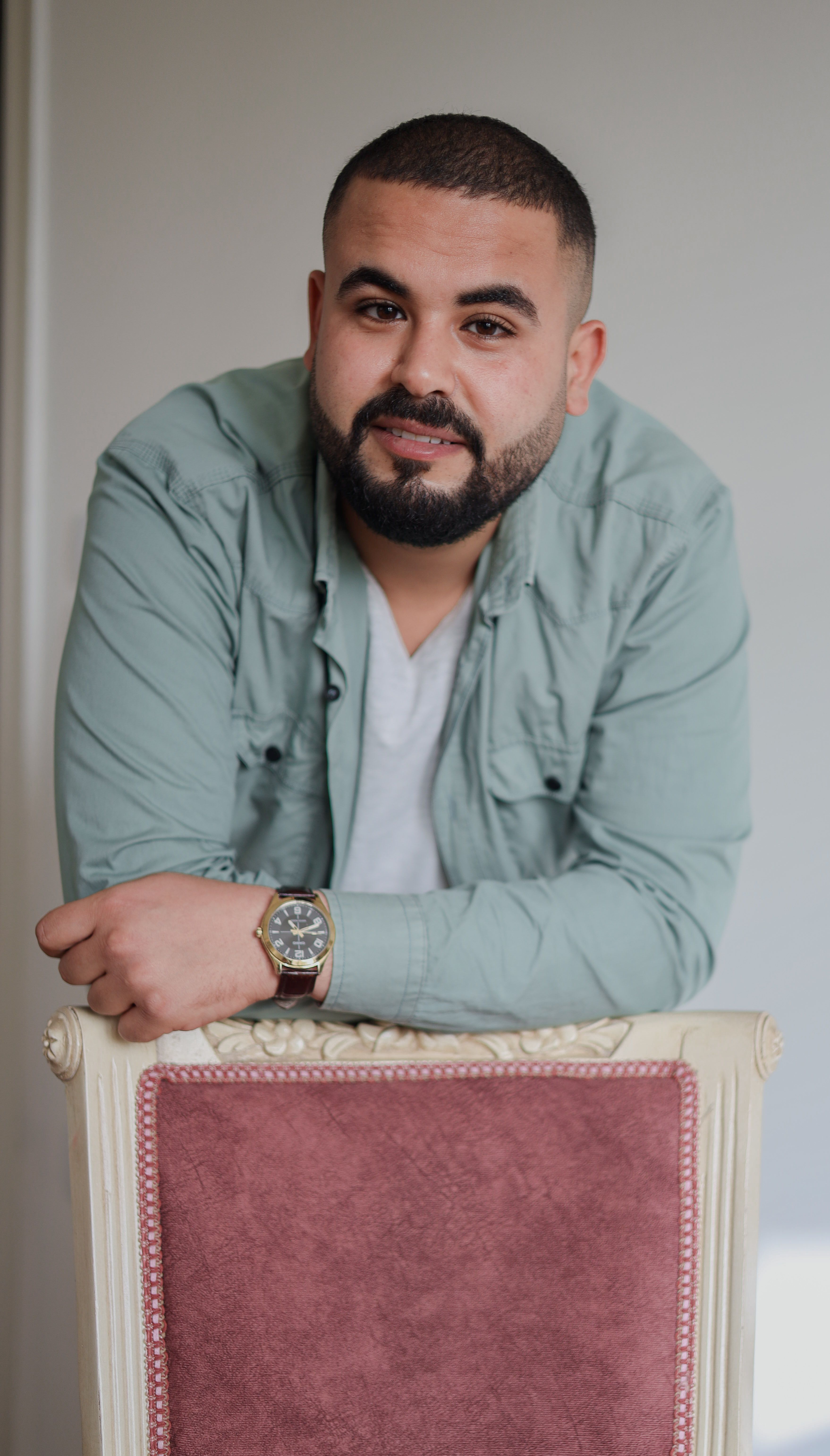 An adult man with a beard and wristwatch leans over a vintage chair indoors.