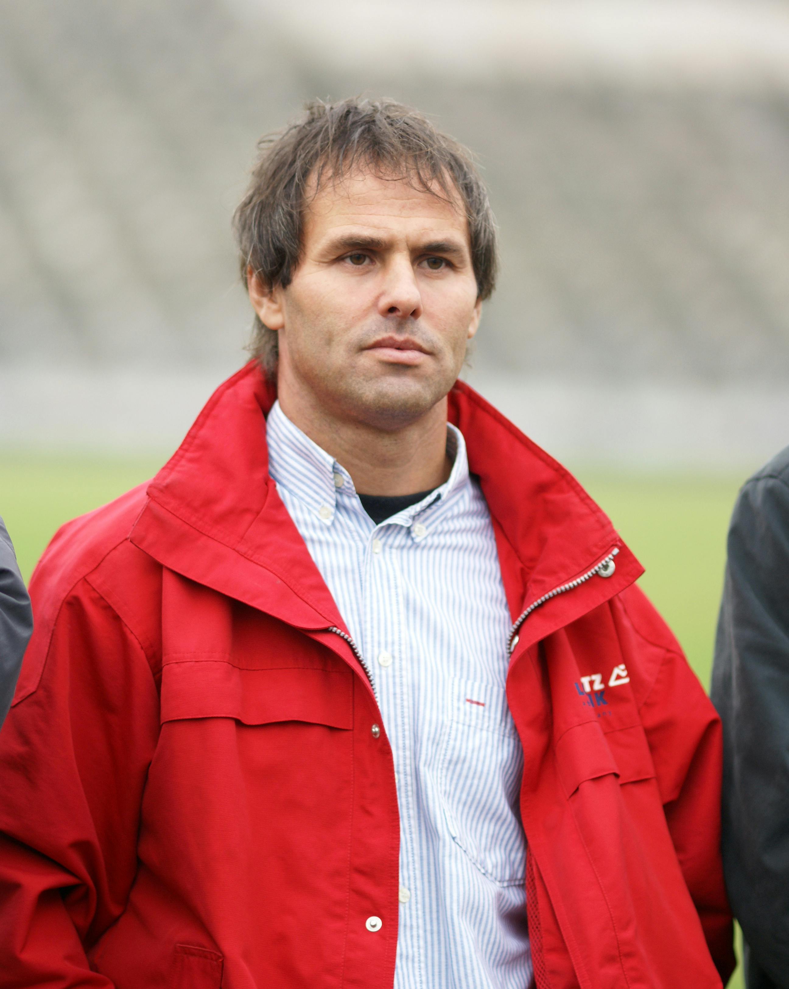 A man in a red jacket standing outdoors in a stadium setting, focused expression.