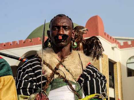 A man in traditional attire poses with an eagle on his shoulder outdoors.