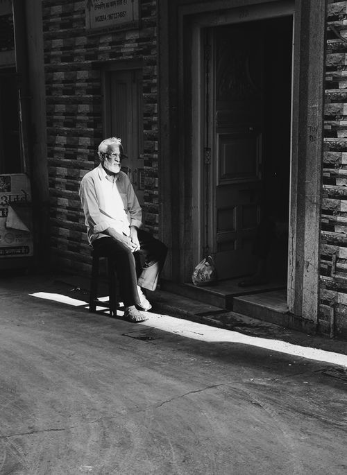 Free Black and white portrait of an elderly man sitting on a stool in a sunlit urban alley. Stock Photo