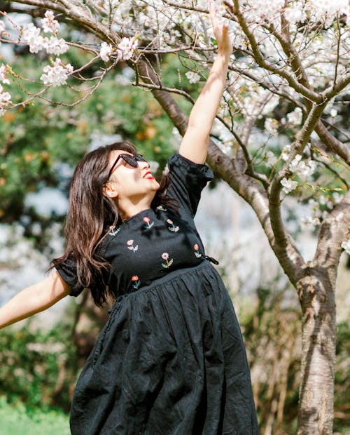 A Woman in a Black Dress Reaching a Cherry Blossom