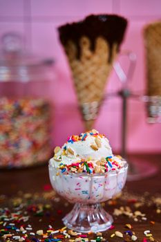 Ice cream sundae topped with vibrant sprinkles and nuts in a glass cup, set against a pink background.