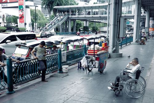 Free Street view in Bangkok showcasing tuk-tuks, a street vendor, and an elderly man in a wheelchair. Stock Photo