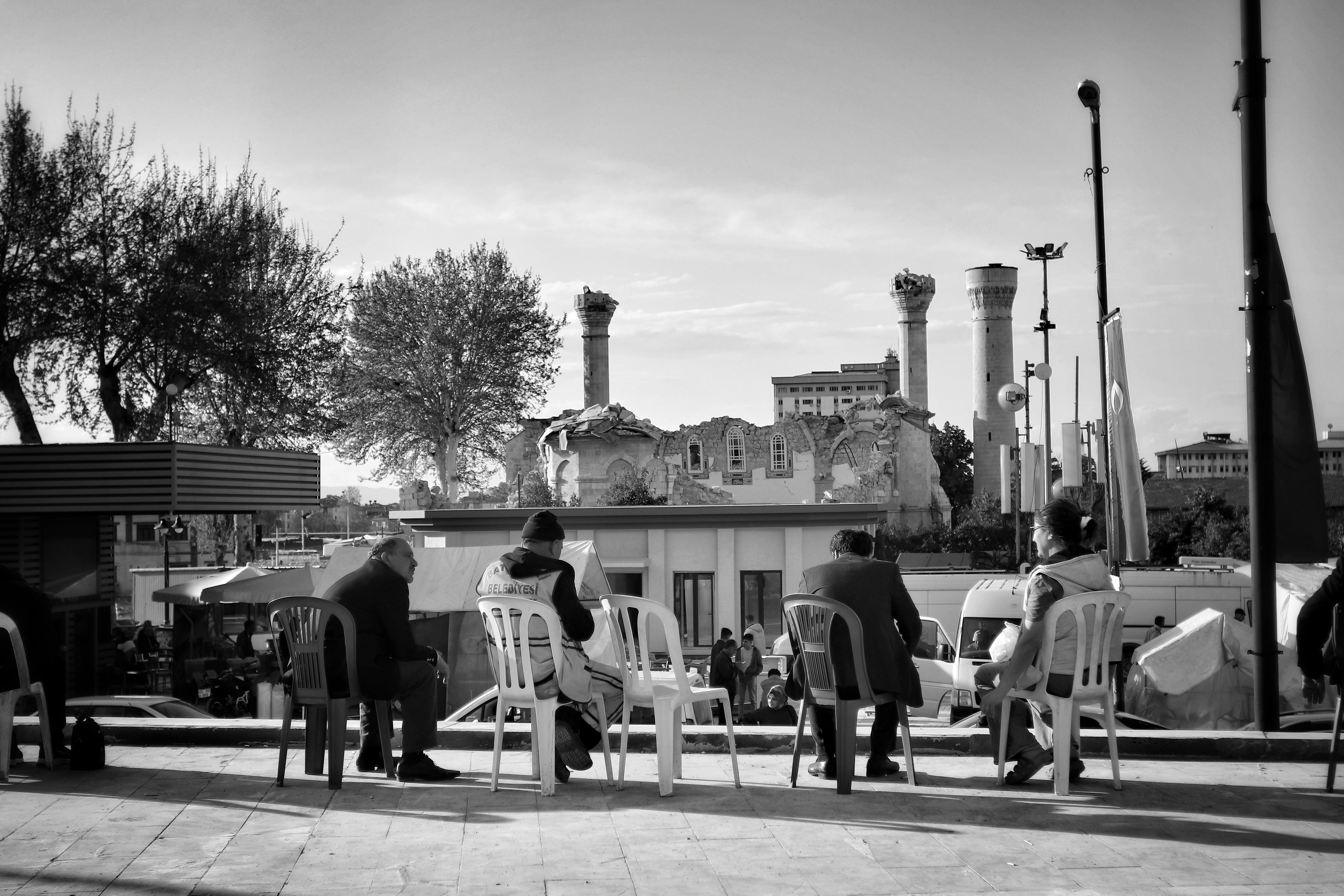 Black and white image capturing people sitting outdoors with ancient ruins in background. (Photo by Serkan  Gönültaş on Pexels)