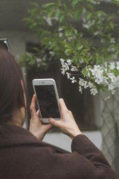 A woman takes a photo of white blossoms with her smartphone, showcasing technology and nature interaction.